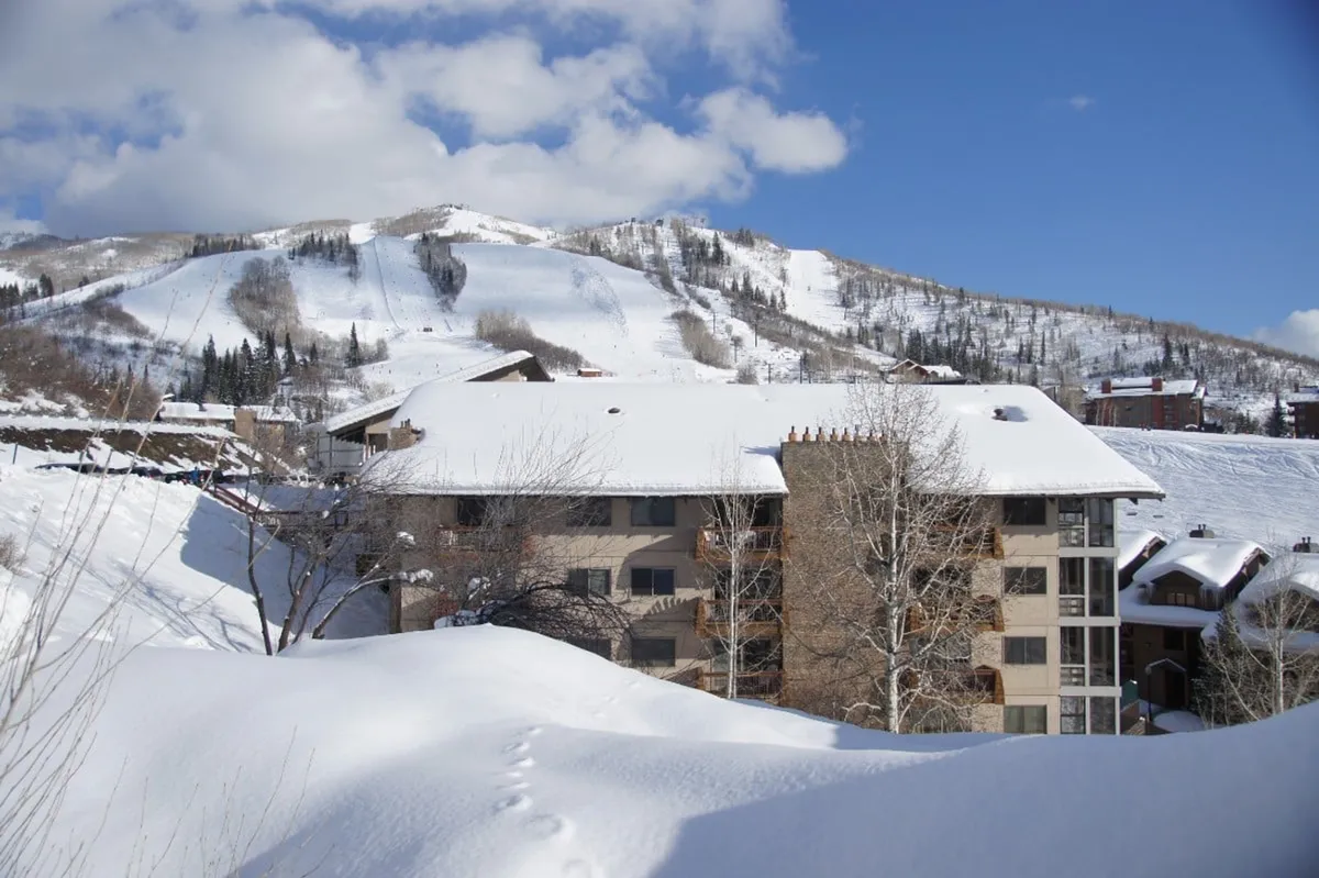 A tranquil exterior shot of snow-covered mountains, the building itself, and bare trees. The scene evokes both relaxation and adventure, with the building positioned as a welcoming retreat after a day spent exploring the base village and slopes.