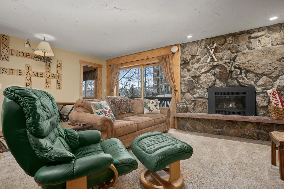 A cozy living room with a stone fireplace as the focal point, a large window offering a glimpse outside, a comfortable sofa, and a green recliner. The room exudes warmth, inviting guests to relax and unwind after a day of outdoor adventure.