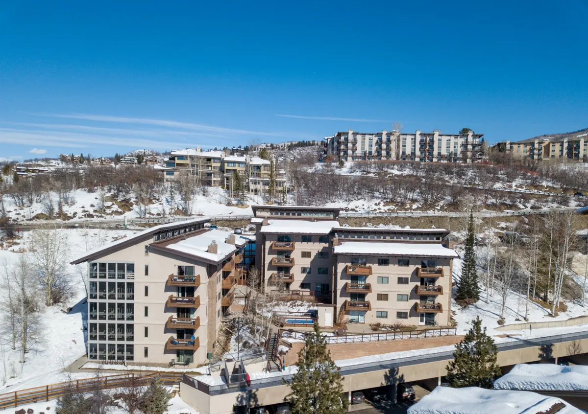 A three-story building with balconies surrounded by a pristine, snow-covered landscape and nearby residential structures. The scene is peaceful but clearly positioned in the heart of mountain adventure territory, with visible trails and easy access to outdoor activities.
