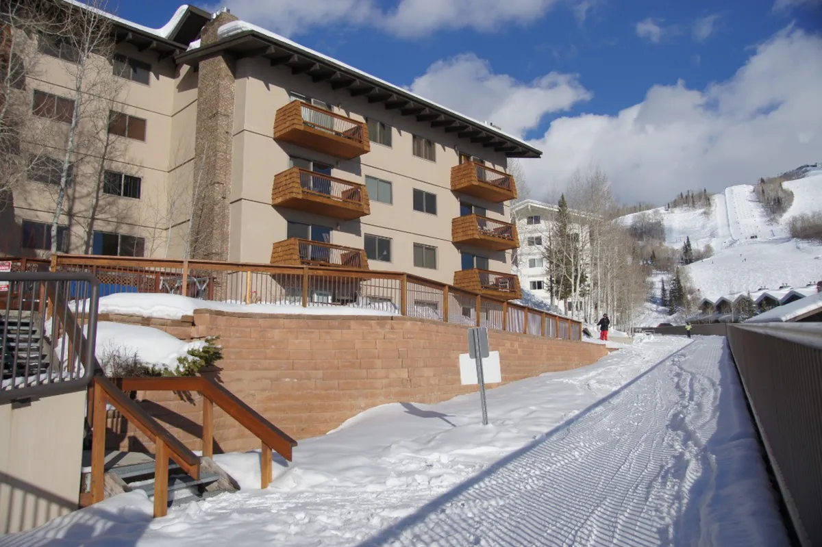 An inviting exterior shot showing wooden balconies along a snow-covered walkway, framed by a striking mountain backdrop. The scene evokes a sense of coziness and accessibility to the outdoors, with visual cues of both adventure and relaxation.