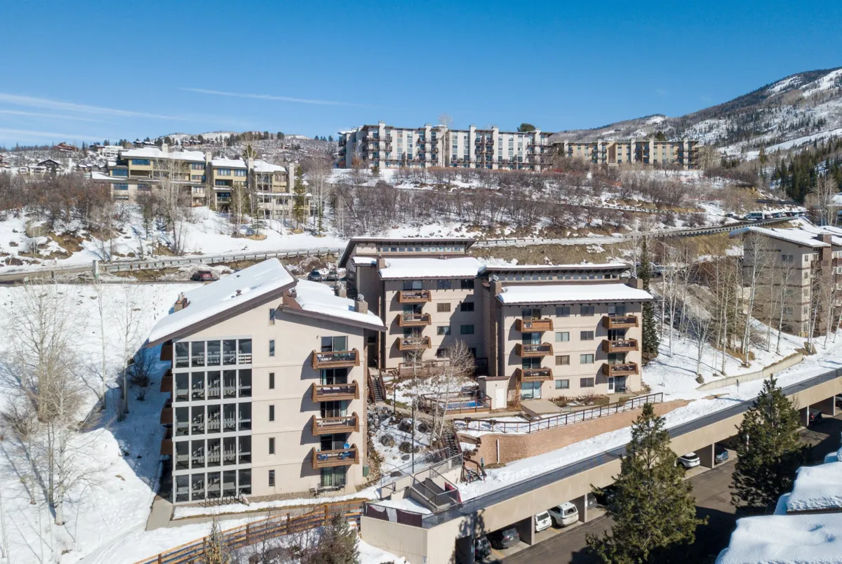 This exterior image captures snow-covered buildings surrounded by trees, with a majestic mountain backdrop and a well-kept sidewalk leading toward the base village area. The inviting scene highlights the easy walkability and scenic atmosphere of Steamboat’s base area.