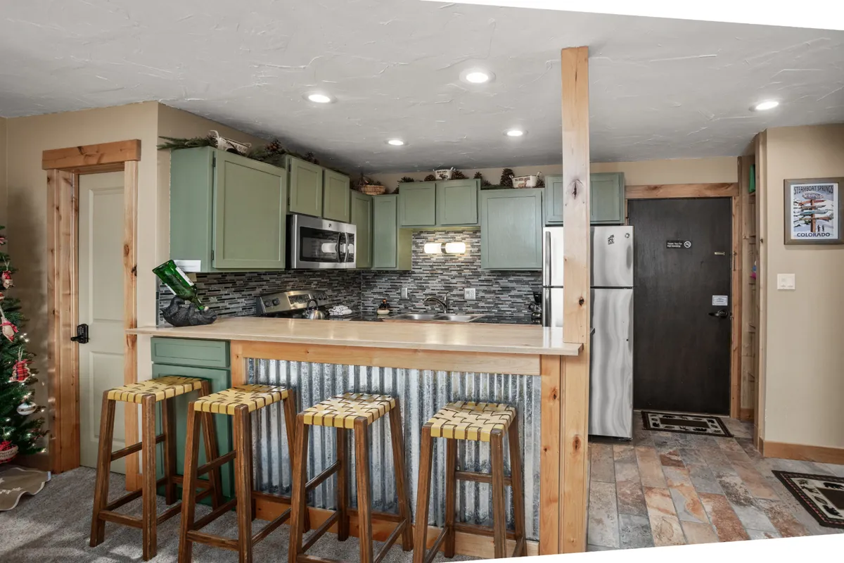 A modern kitchen bar is the focal point, complete with stylish stools, a decorative backsplash, gleaming stainless steel appliances, and a festive Christmas tree in the background. The kitchen is clearly newly renovated, offering a bright and social space for cooking, chatting, and sharing meals.