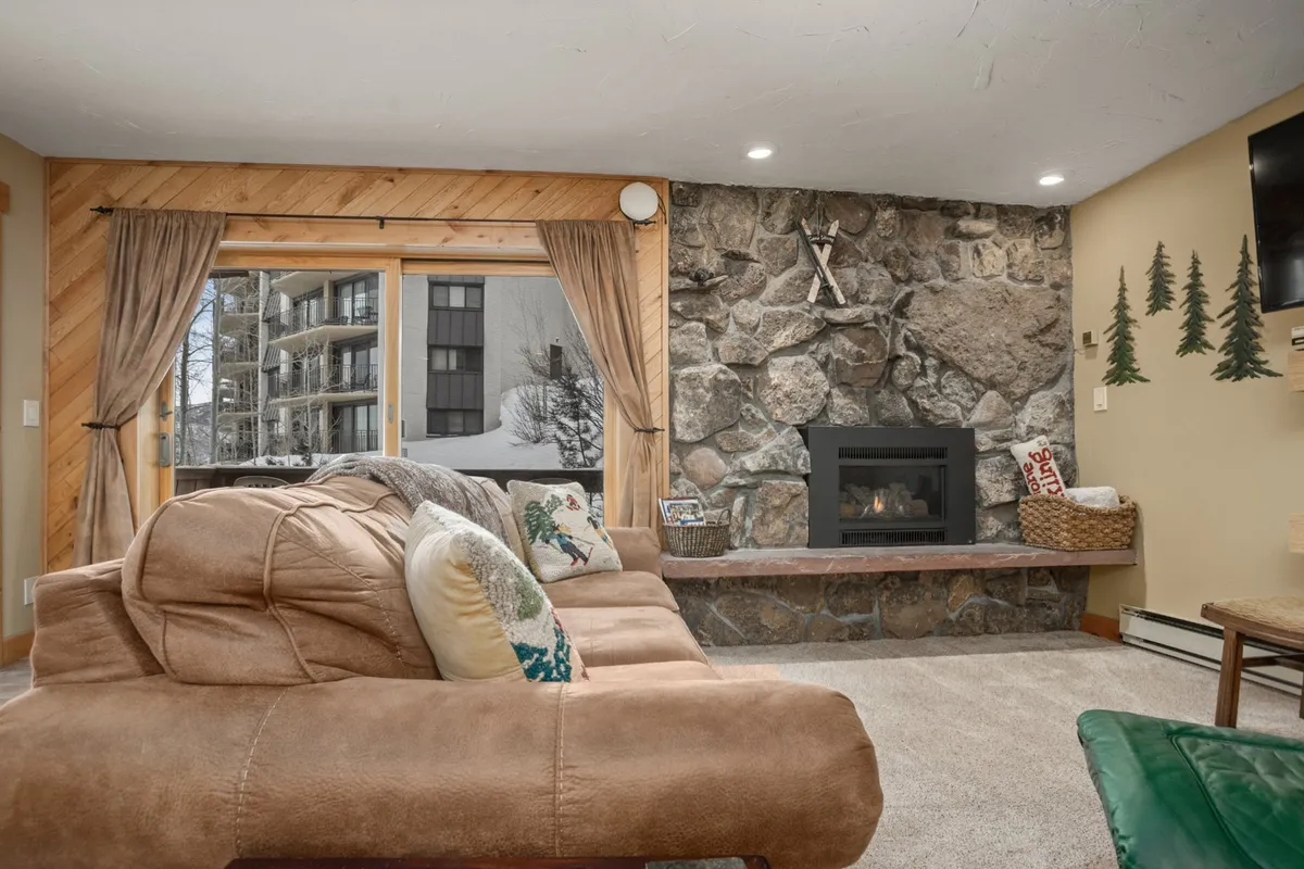 A comfortable living space centered around a stone fireplace, with a large window offering a clear view of the outdoors. There is a plush couch and the ambiance is inviting, ideal for both social gatherings and quiet moments watching snow or wildlife outside.