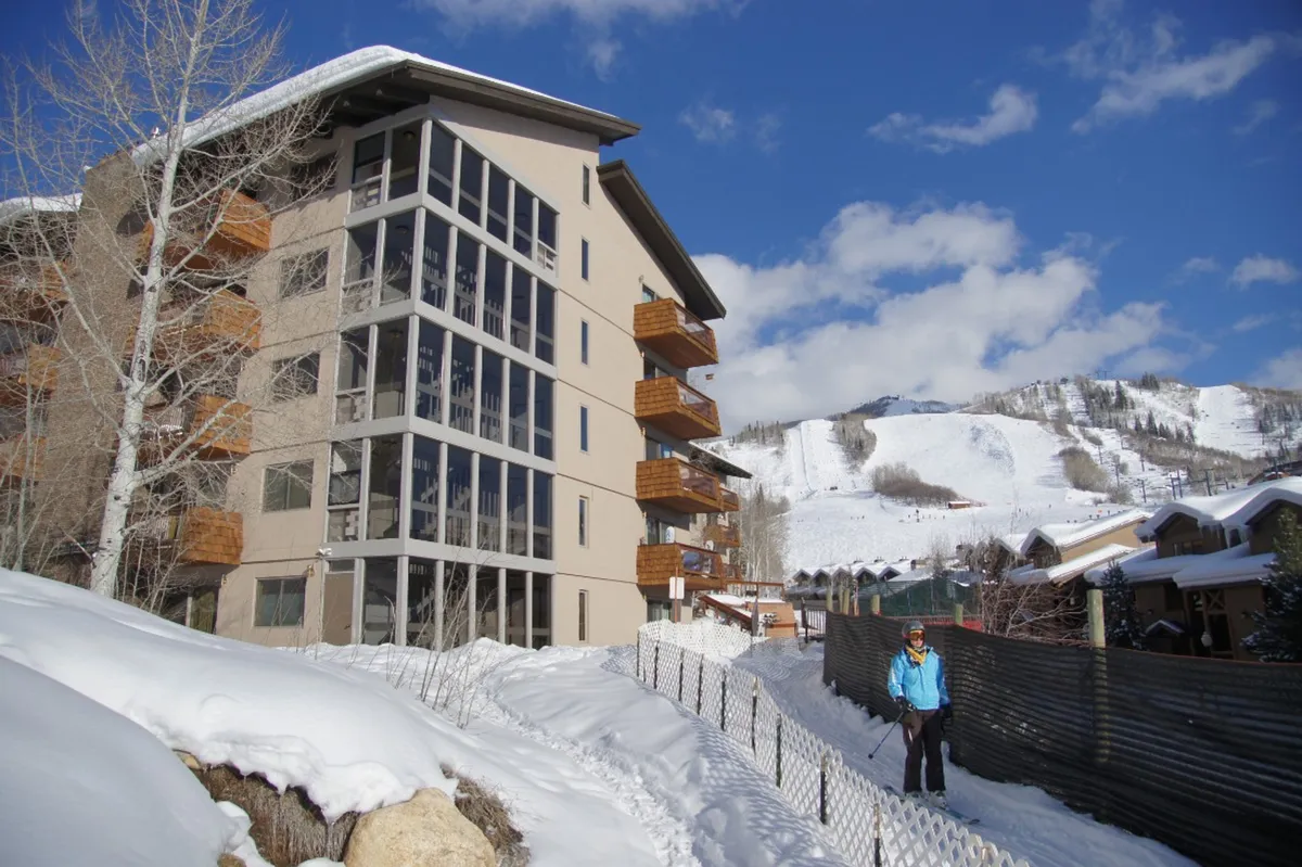 A modern building facade set against a snow-covered landscape, with visible ski slopes and a person in winter attire suggesting active pursuits. The image captures the excitement and accessibility of the slopes, showing both the proximity to skiing and a welcoming home base.