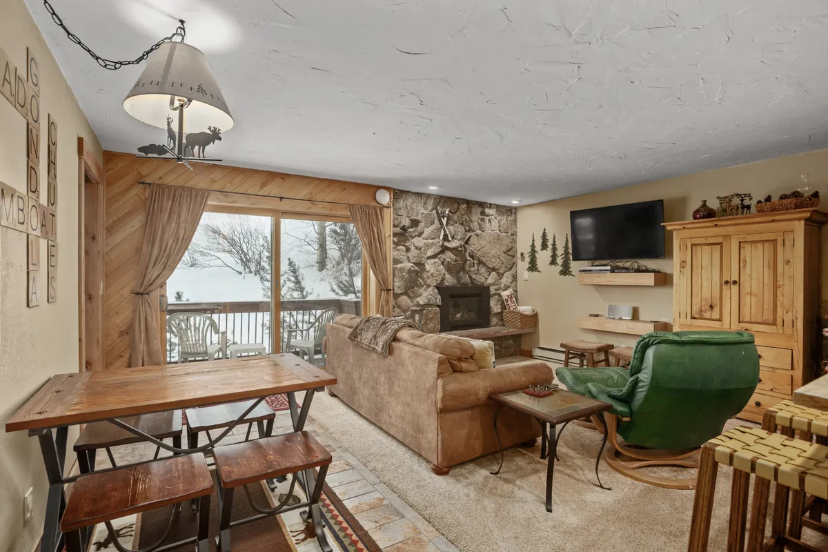 This image captures a warm and welcoming living area featuring a stone fireplace, large windows that frame the view outside, a wooden dining table, and a green armchair. Sunlight floods the space, suggesting a direct connection to the outdoors and the changing seasons.