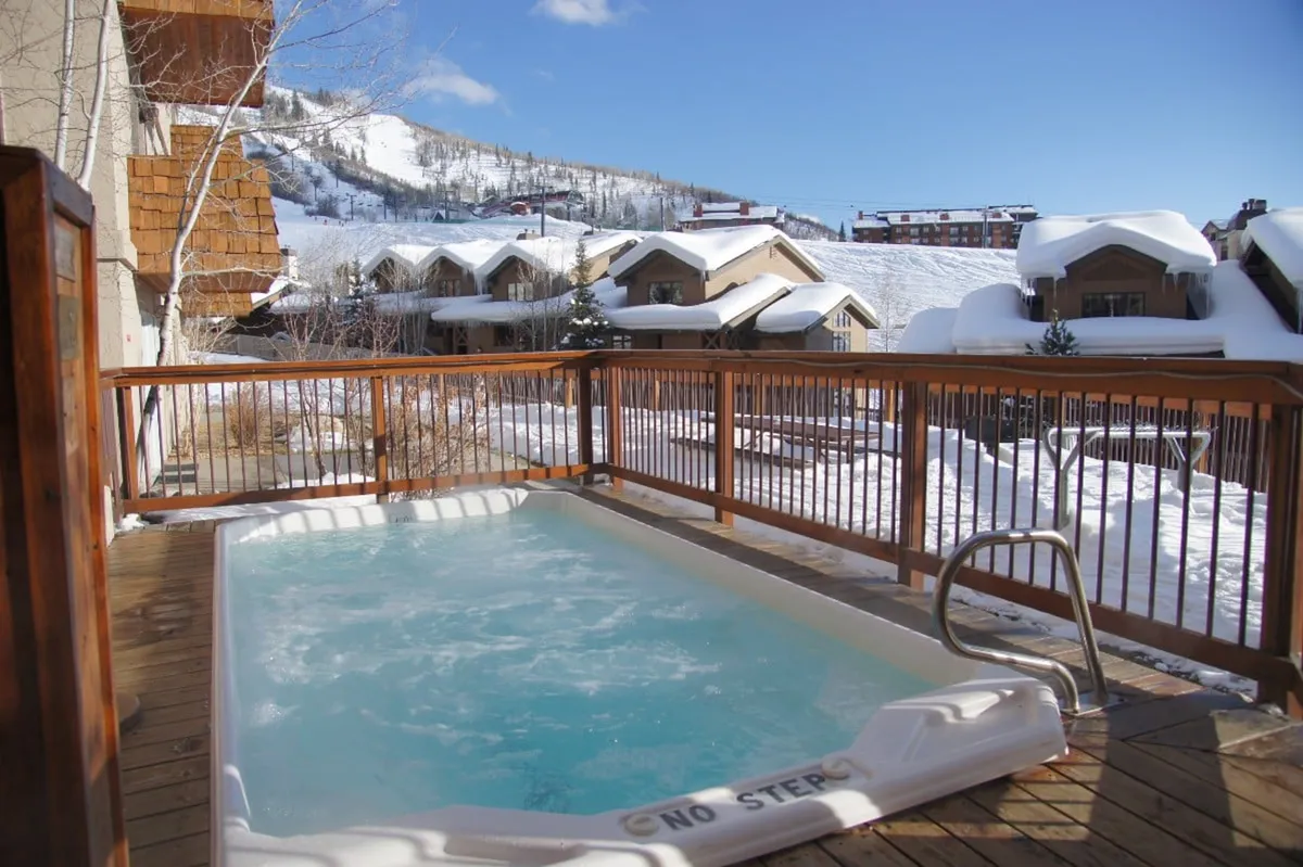 This image features a spacious outdoor hot tub set on a wooden deck, surrounded by a snowy mountain landscape. The hot tub overlooks majestic mountain views, providing a sense of relaxation and immersion in nature. The steam rising from the hot tub contrasts with the crisp, snowy environment, highlighting its appeal after a day of skiing or hiking.