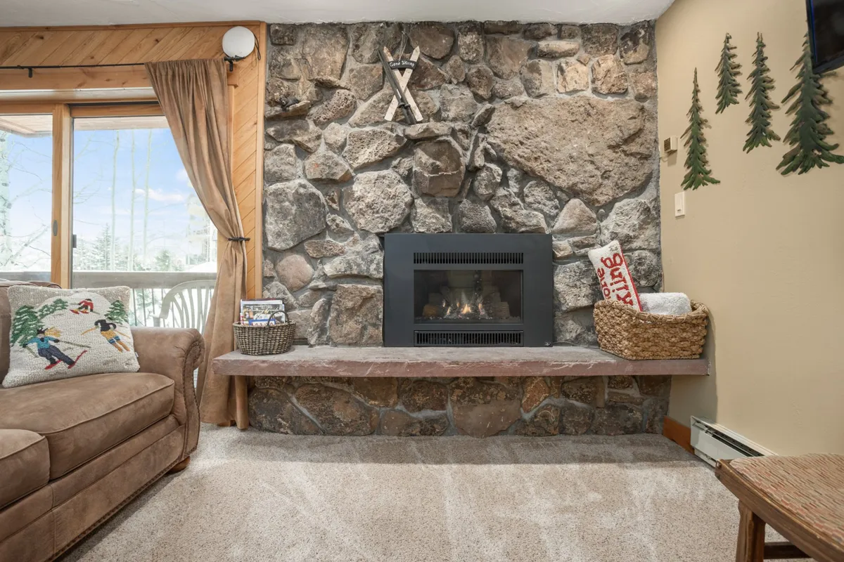 A warm and inviting living space with a stone fireplace, comfortable sofa accented by decorative pillows, and wooden details that create a mountain-lodge atmosphere. The scene evokes the comfort and relaxation available within the condo.