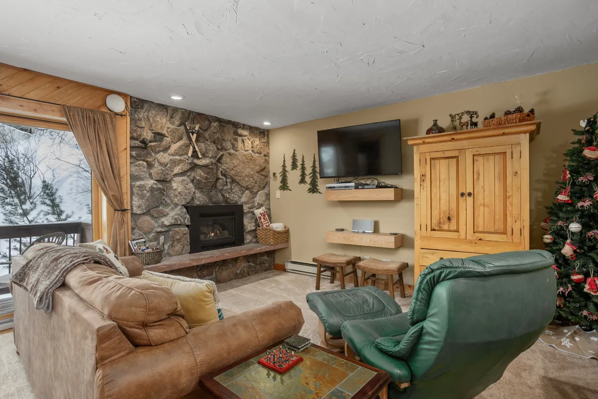 This inviting living room features a striking stone fireplace, wooden furniture, a Christmas tree, and a wall-mounted television. The space is arranged for comfortable gatherings, with plush seating perfect for unwinding after a day outdoors. Festive accents and an expansive window add to the room’s warmth and charm.