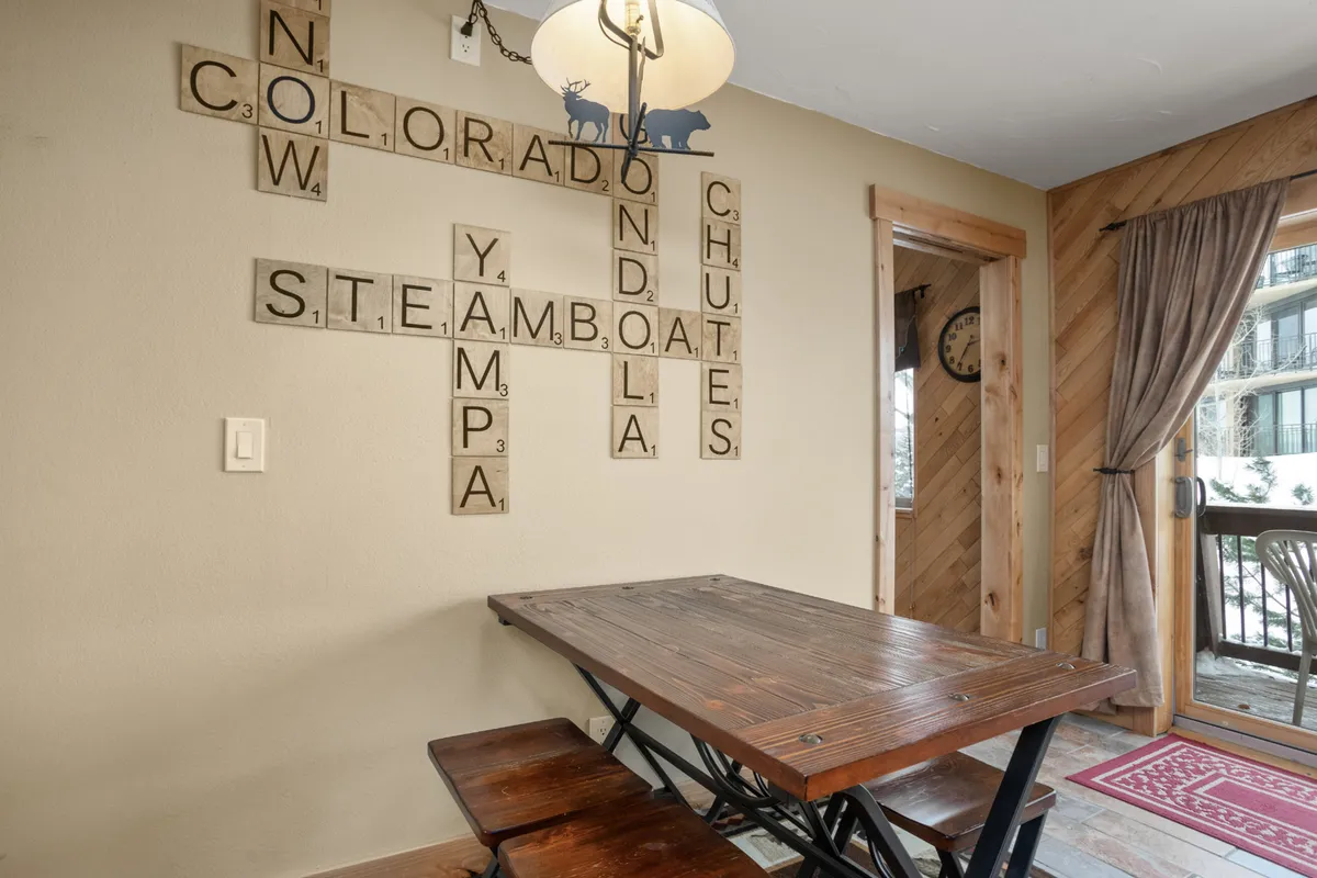 A cozy dining nook is set with a wooden dining table beneath a chic chandelier, flanked by scrabble-style wall art. The setup is intimate and inviting, evoking images of shared breakfasts, dinners, or game nights with family and friends.