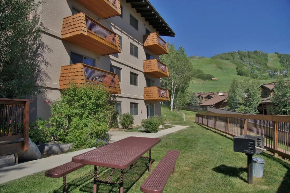 The image showcases the exterior of the condo, featuring inviting balconies with warm wooden accents overlooking a lush green lawn. A picnic table sits in the foreground, surrounded by vibrant greenery, hinting at opportunities for outdoor dining and relaxation against a scenic mountain backdrop.